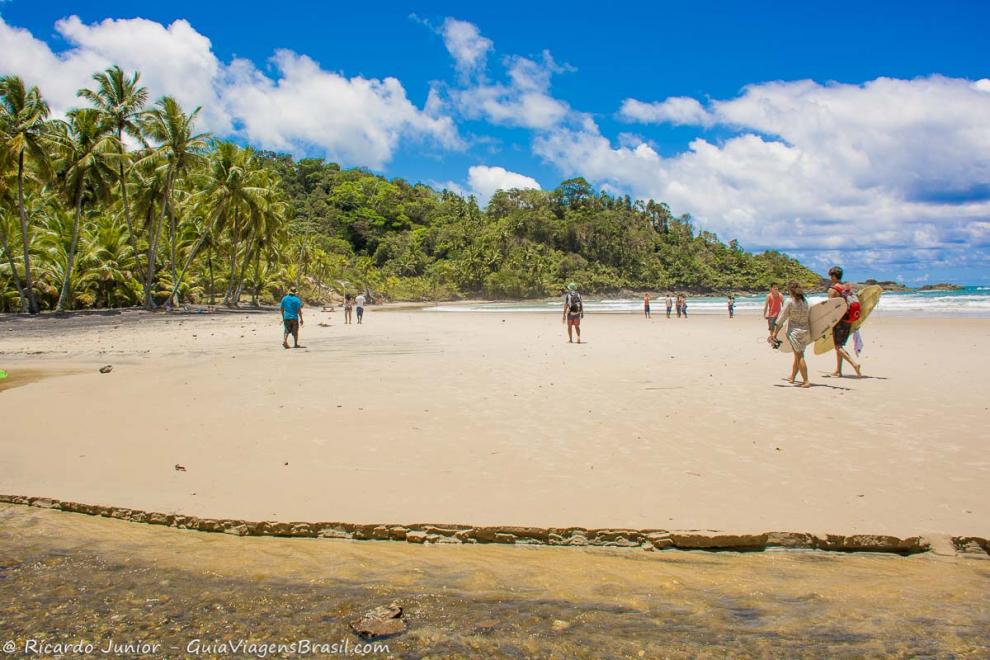 Imagem de turistas com prancha caminhando pelas areias.